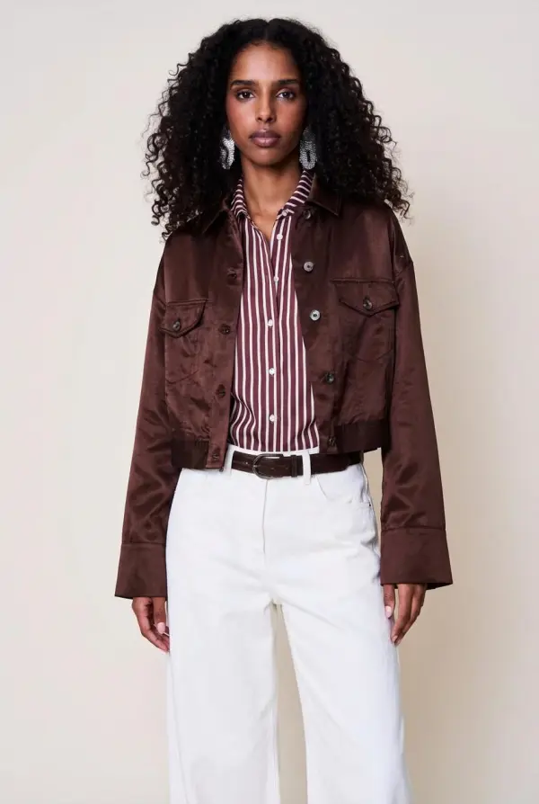 A Woman Is Showcasing A Stylish Outfit Featuring A Striped Shirt A Brown Jacket And White Pants Against A Neutral Background